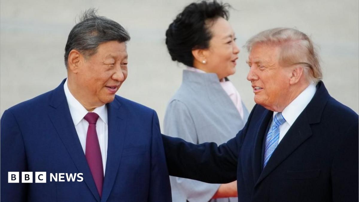 President Xi smiles and looks toward Trump, who has his hand on Xi's shoulder. Both are in suits and their haor is slightly tussled by wind. Xi's wife, in a light blue coat, looks away in the…