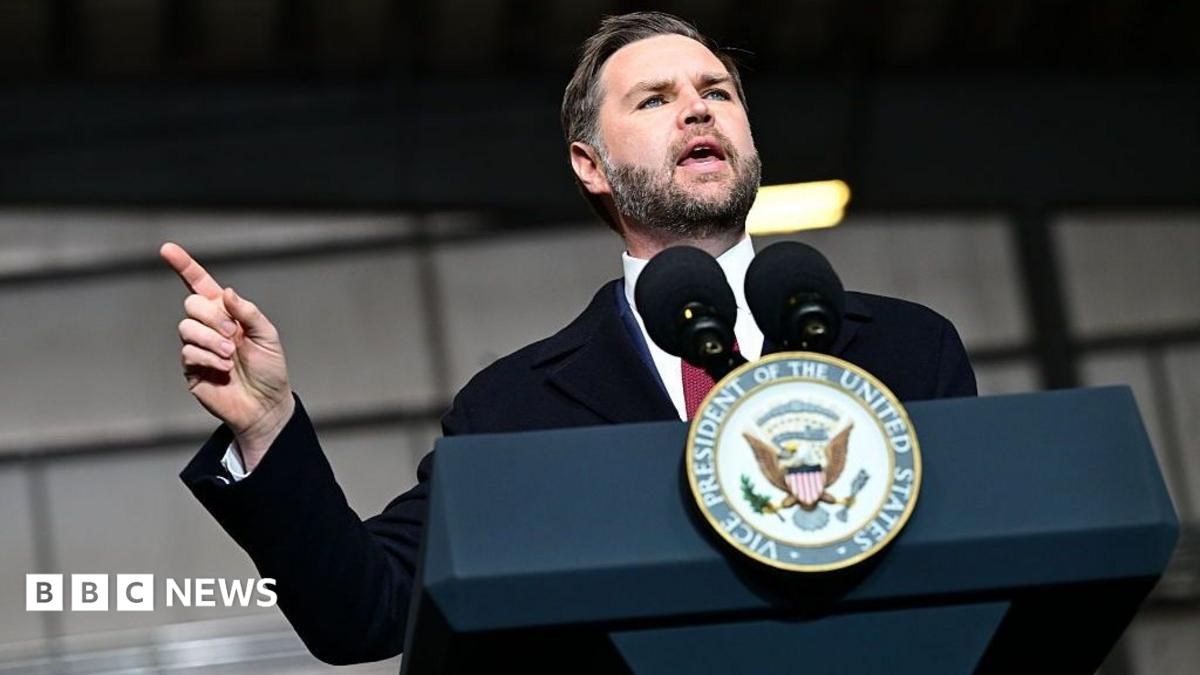 JD Vance stands before a navy blue podium with the vice-presidential seal on it, and two microphones. He is in a suit and tie and is pointing to the right as he gestures